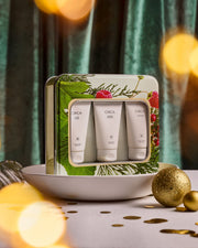 Christmas Hand Care Trio botanical gift tin styled in a bowl with gold baubles and warm festive lights.