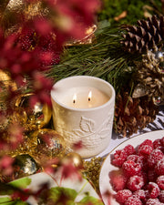 Close-up of a lit Raspberry & Rhubarb candle in its embossed ceramic vessel among festive greenery and raspberries.