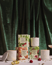 Three festive 300g candles with embossed ceramic vessels, lids and botanical gift boxes against green velvet.